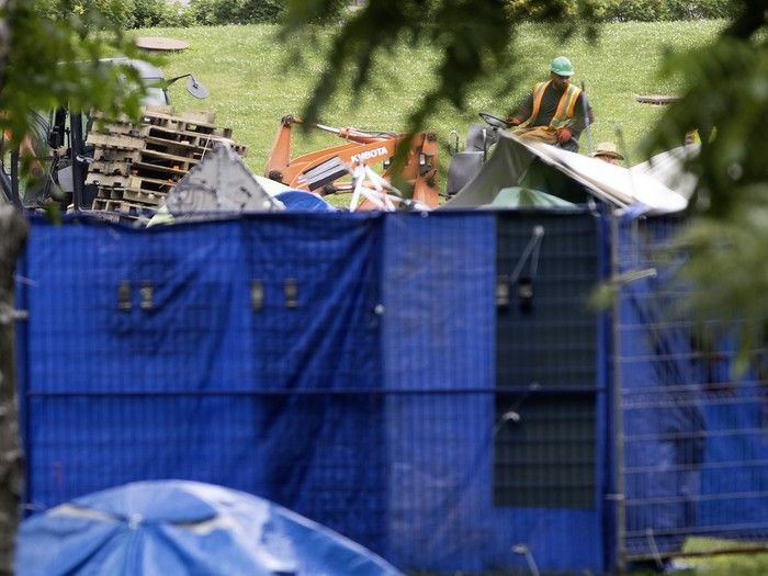 Pallets and tents are seen from behind blue tarp on a fence. There is an excavator in the background.