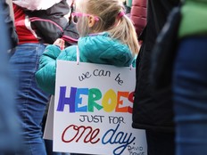 A girl, seen from behind, has a sign on her back that says We Can Be Heroes just For One Day - David Bowie
