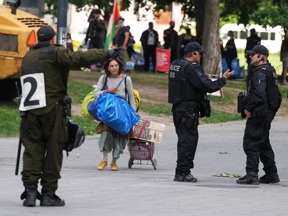 A woman with a shopping cart and bags walks on a street past police officers.