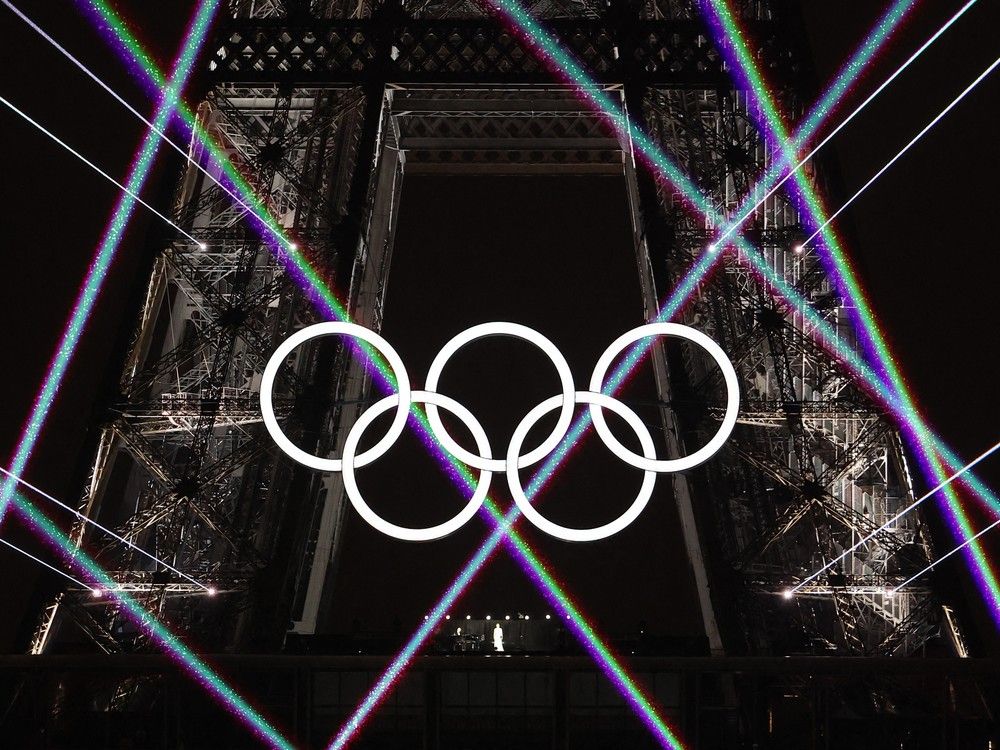 A figure in white appears below the Olympic rings on the Eiffel Tower at night