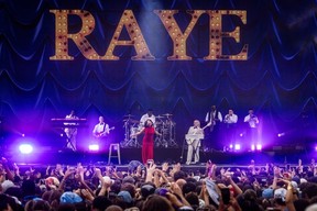 Raye, dressed in red, performs in front of a festival crowd. Behind her is a band in white and a sign with her name.