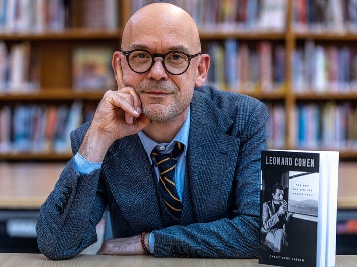 a man in a suit rests his head on his upraised arm as he sits next to a book about leonard cohen.