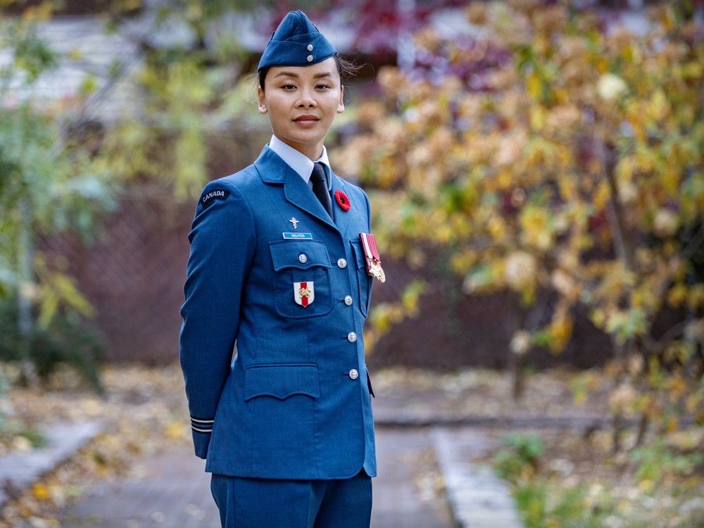 a woman in a blue army uniform poses on a path in what appears to be a park, surrounded by multi-coloured leaves.