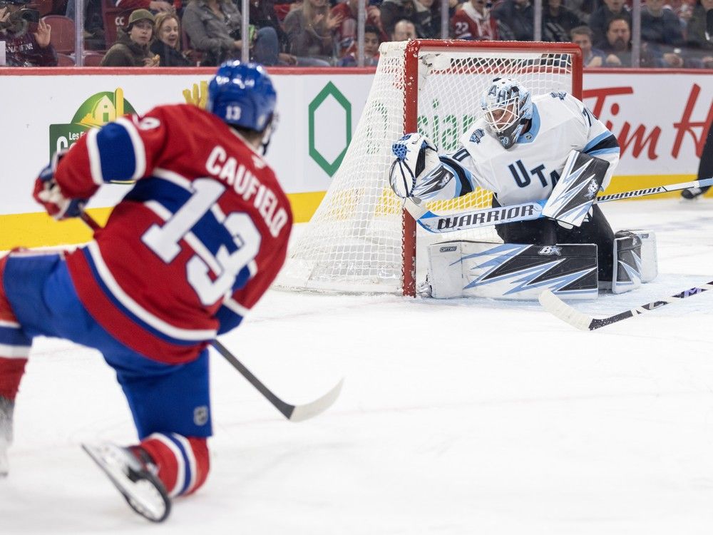 Cole Caufield on one knee following through on a slapshot, while the Utah goalie in the distance extends his glove out to the side