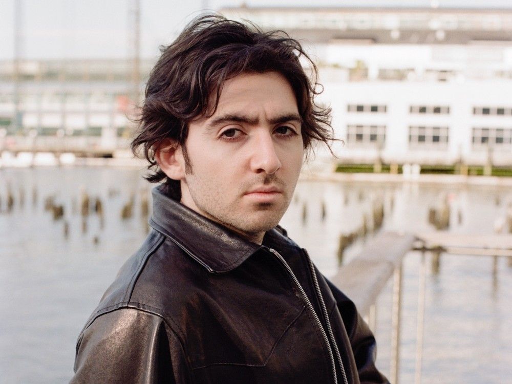 a young man in a leather jacket stares at the camera in an outdoor setting.