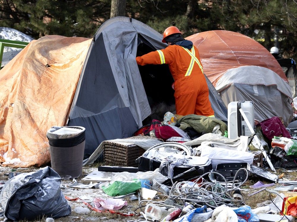 a worker in orange gear looks into a tent made of tarps. there is debris surrounding it.