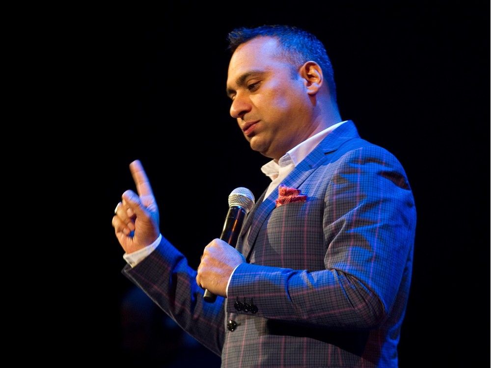 comedian russell peters makes a face while raising a finger on stage.