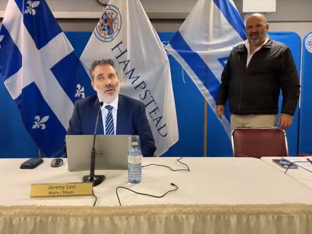 jeremy levi sits at a table with a nameplate and microphone in front of him and quebec, hampstead and israel flags behind him, while jason farber stands behind a chair next to him