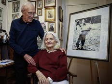 An older couple pose in a room of vintage family photos. The wife is seated while the husband looks affectionately at her.