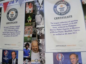 Philippos Stavrou Platini, owner of a museum poses for a photo by posters of French former soccer great and former UEFA president Michel Platini and two certificates of Guinness world records at his museum in Mosfiloti village, in the island of Cyprus, Friday, Sept. 8, 2017. A Cypriot man who idolizes Platini has managed clinched two entries in the latest annual Guinness Book of World Records book for his stash of photos, jerseys, shoes, balls and more centered on him. (AP Photo/Petros Karadjias)