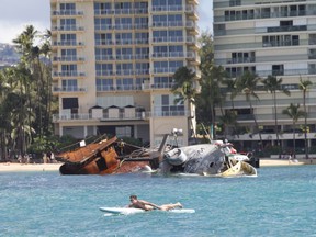 In this Nov. 13, 2017 photo, a man on a surfboard swims past the Pacific Paradise fishing vessel off Waikiki in Honolulu. The U.S. Coast Guard says the fishing vessel that ran aground just off Waikiki more than a month ago is being prepared for removal, possibly by the end of the week. A helicopter hoisted 16 sheets of metal from the boat Sunday, Nov. 19, in a bid to remove weight so it can be floated again. (AP Photo/Marco Garcia)