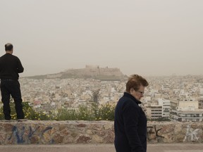 A woman walks as a in the background is seen the city of Athens with the Acropolis hill covered with dust from the Saharan desert, on Monday, March 26, 2018. Swathes of Greece have been shrouded in haze as persistent southerly winds have carried waves of dust from the African continent across the Mediterranean.