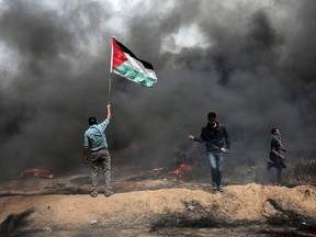 A Palestinian protester waves the national flag as smoke billows from a tire fire during clashes with Israeli forces on April 20, 2018, east of Khan Yunis, in the southern Gaza Strip during mass protests along the border of the Palestinian enclave, dubbed "The Great March of Return," which has the backing of Gaza's Islamist rulers Hamas.