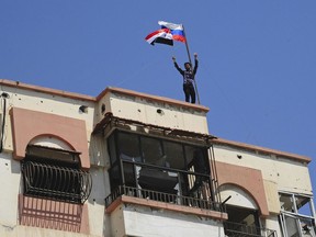 This photo released Saturday, April 14, 2018, by the Syrian official news agency SANA, shows a man giving the victory sign next to Syrian and Russian flags on  top of a damaged apartment, after Syrian police units entered the town of Douma, the site of a suspected chemical weapons attack and the last rebel-held town in the eastern Ghouta, near Damascus, Syria. Syrian state TV is broadcasting the deployment of 5,000 policemen and internal security in Douma where an alleged chemical attack last weekend triggered unprecedented joint US, British, French strikes in Syria. (SANA via AP)