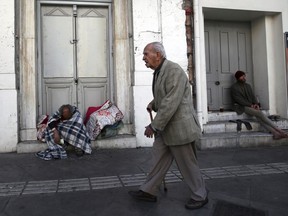 An elderly man passes by homeless men in Athens, Monday, April 23, 2018. Greece has beaten its bailout budget targets for a third successive year and eased its massive debt burden by a fraction as the country prepares to exit its international rescue program in four months.