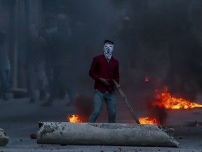 A masked Kashmiri protester watches from a distance as Indian policemen fire tear gas shells towards him during a protest in Srinagar, Indian controlled Kashmir, Thursday, June 21, 2018. Shops, businesses and schools shut down in the Indian portion of Kashmir Thursday in response to a strike call given by separatists against recent civilian deaths and the killing of a senior journalist Shujaat Bukhari.