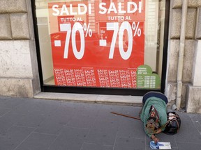 A woman begs for money near a shop window promoting sales up to 70 per cent discount, in downtown Rome, Wednesday, July 25, 2018.