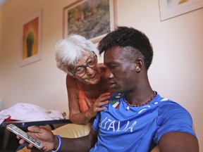 In this photo taken on Sunday, July 15, 2018, asylum seeker from Mali, Moriba Mamadou Diarra, 18, looks at his mobile phone as Barbara Di Clemente, 79-year-old Italian grandmother, talks to him at her home in Rome.  Di Clemente has been hosting Diarra in her home for a few months through a program called "Refugees Welcome" where Italian families can apply to open their homes and host young refugees in need.