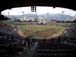 In this Oct. 12, 2018 photo, baseball players and fans stand for the national anthem during the opening season baseball game between Leones de Caracas and Tiburones de la Guaira in Caracas, Venezuela. For the second straight year, state-run oil company PDVSA had to step in with a $12 million lifeline to pay for everything from imported baseballs to the salaries of seven foreign-born players _ most of them minor league prospects from the U.S. _ on each team's roster.