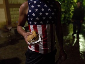 A Honduran migrant wearing a U.S. flag motif shirt, holds a sandwich at an improvised shelter in Chiquimula, Guatemala, Tuesday, Oct. 16, 2018. U.S. President Donald Trump threatened on Tuesday to cut aid to Honduras if it doesn't stop the impromptu caravan of migrants, but it remains unclear if governments in the region can summon the political will to physically halt the determined border-crossers.