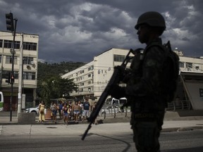 In this May 19, 2018 photo, a soldier patrols during an operation while pedestrians wait to cross the street in the Bateau Mouche slum of Rio de Janeiro, Brazil. This particular street separates the area controlled by drug traffickers in the Bateau Mouche slum, from the area controlled by a militia in the Chacrinha slum.