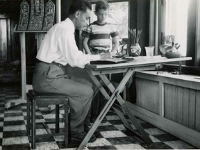 Black and white photograph depicting artist Vincenzo Poggi working at his desk in front of a window at his studio in Montreal, Quebec. His young son Maurice Poggi is standing beside his father observing him work. In the left background a model for a stained glass window is visible sitting on an easel.