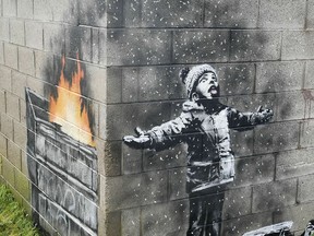 An artwork by Banksy on the side of a garage depicts a child dressed for snow playing in the falling ash and smoke from a skip fire, in Port Talbot, Wales.