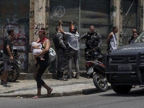 A woman carries a baby as she walks past police searching several men, during an operation targeting drug traffickers in the Santa Teresa neighborhood of Rio de Janeiro, Brazil, Friday, Feb. 8, 2019. Law enforcement officials in Brazil's second largest city say that at least 11 suspected drug traffickers were killed in a shootout with police in a slum located in the bohemian Santa Teresa neighborhood.