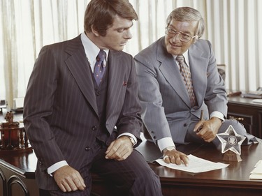 Two businessmen of the '70s, with bold patterned suits, thick sideburns, patterned ties and bold cuffs.