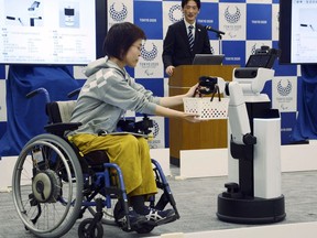 A robot passes a basket containing drinks to a woman in wheelchair during an unveiling event in Tokyo Friday, March 15, 2019. Organizers on Friday showed off robots that will be used at the new National Stadium to provide assistance for fans using wheelchairs. (Kyodo News via AP)