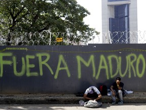 A man reaches into his bag to retrieve some food waste that was given to him by a Chinese restaurant as he sits in front of security fence spray-painted with a message that reads in Spanish: "Maduro out," in Caracas, Venezuela, Wednesday, March 20, 2019. U.S. President Donald Trump said Tuesday that the United States could impose harsher sanctions on Venezuela in its campaign to oust Maduro.