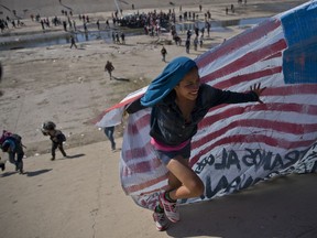 FILE - In this Nov. 25, 2018 file photo, a migrant woman helps carry a handmade U.S. flag up the riverbank at the Mexico-U.S. border after getting past Mexican police at the Chaparral border crossing in Tijuana, Mexico, as a group of migrants tries to reach the U.S.  Activists, officials and social workers in Central America were staggered by the idea that U.S. President Donald Trump thinks he will help reduce immigration, by cutting off nearly $500 million in aid to Honduras, Guatemala and El Salvador; exactly the opposite will happen, they say.
