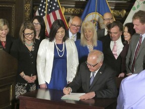 Jenny Teeson, center in white, of Andover, Minnesota, looks on as Minnesota Gov. Tim Walz signs a bill at the Capitol in St. Paul, on Thursday, May 2, 2019, repealing a Minnesota law that prevented prosecutors from filing sexual assault charges against people accused of raping their spouse. Teeson, testified before legislative committees earlier this year about how her now ex-husband drugged her and made a video of himself raping her while she was unconscious. Prosecutors dropped rape charges because of the old law, and he served just 30 days in jail for invasion of privacy.