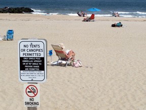 In this Monday, May 20, 2019 photo, beachgoers sit on the sand beyond a sign indicating that smoking is prohibited on the beach in Spring Lake, N.J. A statewide smoking ban is in effect at New Jersey's beaches for the start of the 2019 summer season, although towns can set aside up to 15% as smoking sections.