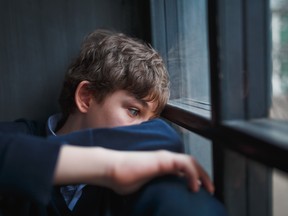 Pensive sad boy teenager in a blue shirt and jeans sitting at the window and closes his face with his hands.