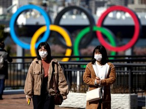 People wearing protective face masks walk in front of the Giant Olympic rings in Tokyo on Feb. 27.