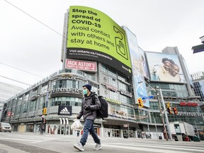 A pedestrian wearing a mask walks across Yonge Street at Dundas Street with a “Stop The Spread Of Covid 19” notice in the background, April 3, 2020.