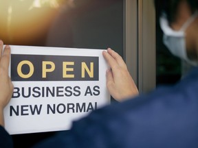 Reopening for business adapt to new normal in the novel Coronavirus COVID-19 pandemic. Rear view of business owner wearing medical mask placing open sign "OPEN BUSINESS AS NEW NORMAL" on front door.