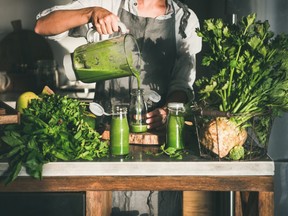 Making green detox take-away smoothie. Woman in linen apron pouring green smoothie drink from blender to bottle surrounded with vegetables and greens. Healthy, weight loss food concept