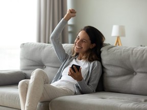 Hilarious euphoric millennial girl getting good news notification via mobile phone, making yes gesture, celebrating success, online lottery win, cheap flight tickets purchase, sitting on couch.