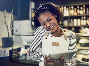 Smiling young African entrepreneur standing at the counter of her cafe talking on a cellphone and using a tablet