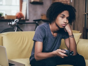 Young African man holding remote control and looking bored while watching TV on the couch at home.