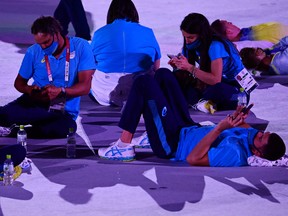 Athletes sit on the ground and look at their phones as delegations enter the Olympic Stadium during the opening ceremony of the Tokyo 2020 Olympic Games.