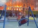 A woman is seen swinging on a play set with a child on her knees as they watch the fire blaze on.