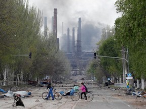 People walk their bikes across the street as smoke rises above a plant of Azovstal Iron and Steel Works during Ukraine-Russia conflict in the southern port city of Mariupol, Ukraine May 2, 2022.