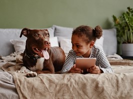 Front view portrait of cute African-American girl lying on bed with big pet dog and smiling, copy space