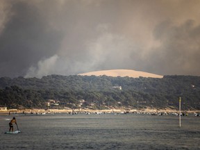 A paddle surfer is seen as smoke laden with ashes coming from a giant wildfire consuming the thousand-year-old forest bordering the Dune du Pilat rises over the beach of Pilat sur Mer, southwestern France, Monday July 18, 2022. France scrambled more water-bombing planes and hundreds more firefighters to combat spreading wildfires that were being fed Monday by hot swirling winds from a searing heat wave broiling much of Europe.