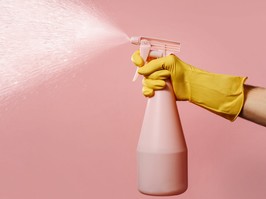 A hand in a silicone household glove holds a liquid spray bottle on a pink background with space for text.. A stream of water hits from the atomizer. The concept of maintaining cleanliness and order in the house.
