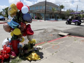 A woman driving a golf cart crosses 33rd Street at Avenue R in Galveston, Texas on Monday, Aug. 8, 2022, in Galveston, Texas, where balloons, flowers and stuffed animals have been left in memory of the victims of a fatal crash involving a golf cart and two automobiles. The four people killed in a weekend golf cart collision in Texas were a grandfather, two of his grandchildren and a niece who were visiting Galveston for a quick vacation before school began, police said.