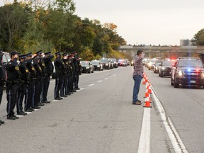 Ontario Provincial Police officer Adam McCourt, left in plainclothes, and Sgt. Jonathan Bouchard, obscured, salute the arriving procession carrying the bodies of two South Simcoe Police officers who were killed responding to a call, in Innisfil, Ont., Friday, Oct. 14, 2022. McCourt served with South Simcoe police and was trained by Morgan Russell, one of the fallen officers.